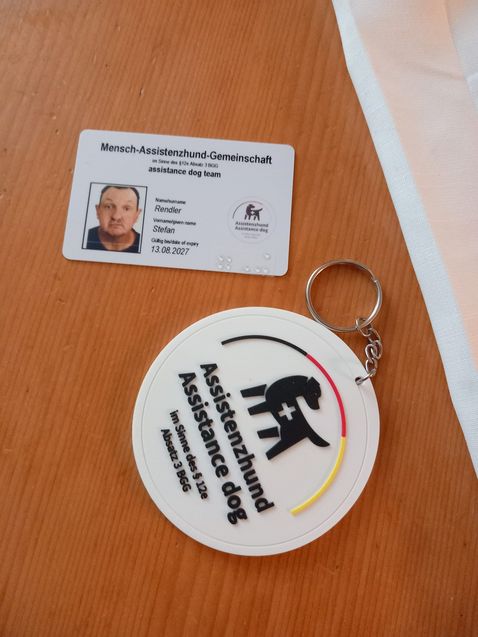 Das Bild zeigt zwei Gegenstände auf einer Holzoberfläche:  1. Eine Identifikationskarte für ein "Mensch-Assistenzhund-Gemeinschaft" Team (auf Englisch "assistance dog team"). Die Karte enthält folgende Informationen: - Ein Passfoto eines Mannes - Name: Rendler - Vorname: Stefan - Gültig bis: 13.08.2027 - Ein Logo eines Hundes mit einem Menschen  2. Ein runder weißer Schlüsselanhänger mit folgenden Elementen: - Die Aufschrift "Assistenzhund" in schwarzen Buchstaben - Ein stilisiertes Bild eines Hundes mit einem Kreuz - Der Text "Assistance dog" in kleinerer Schrift - Ein kleiner Streifen in den Farben Schwarz, Rot und Gelb am unteren Rand  Beide Gegenstände scheinen Teil einer Ausrüstung für Menschen mit Assistenzhunden zu sein, möglicherweise zur Identifikation und Information in der Öffentlichkeit.